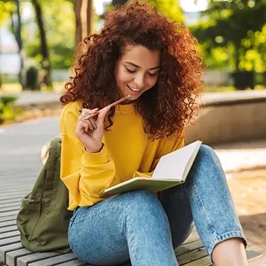 Young beautiful curly student girl sitting outdoors in nature park writing notes in notebook.